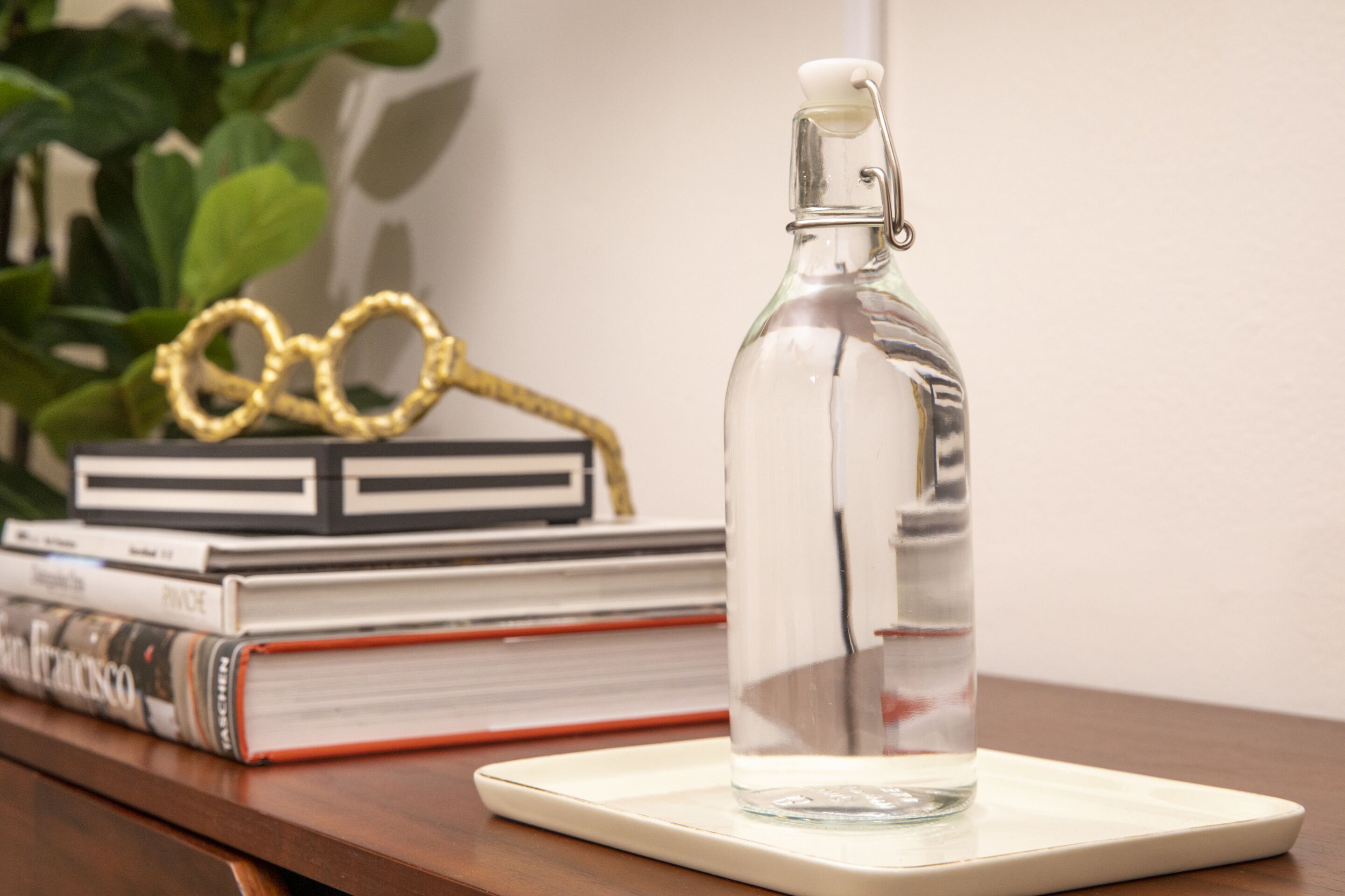 A clear glass bottle with a metal cap on a white coaster, placed on a wooden surface with books and a pair of gold-rimmed glasses in the background.