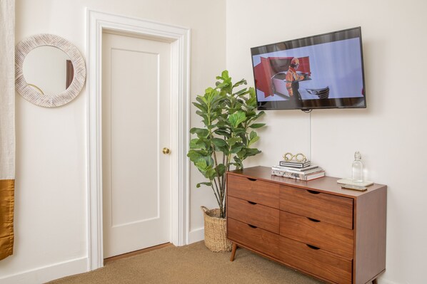 A room with a white door, a round mirror, a wooden dresser, a television on the wall, and a potted plant.