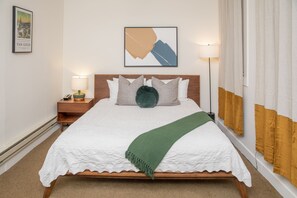 A neatly made bed with a white duvet and a green throw, a wooden headboard, a nightstand with a lamp, and a framed picture on the wall.