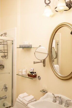 A bathroom with a glass shower enclosure, a sink with two faucets, a large mirror, and wall-mounted shelves with toiletries.