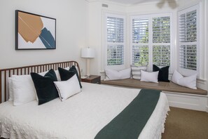 A hotel room with a large bed, a bench with cushions, a window with blinds, and a framed picture on the wall.