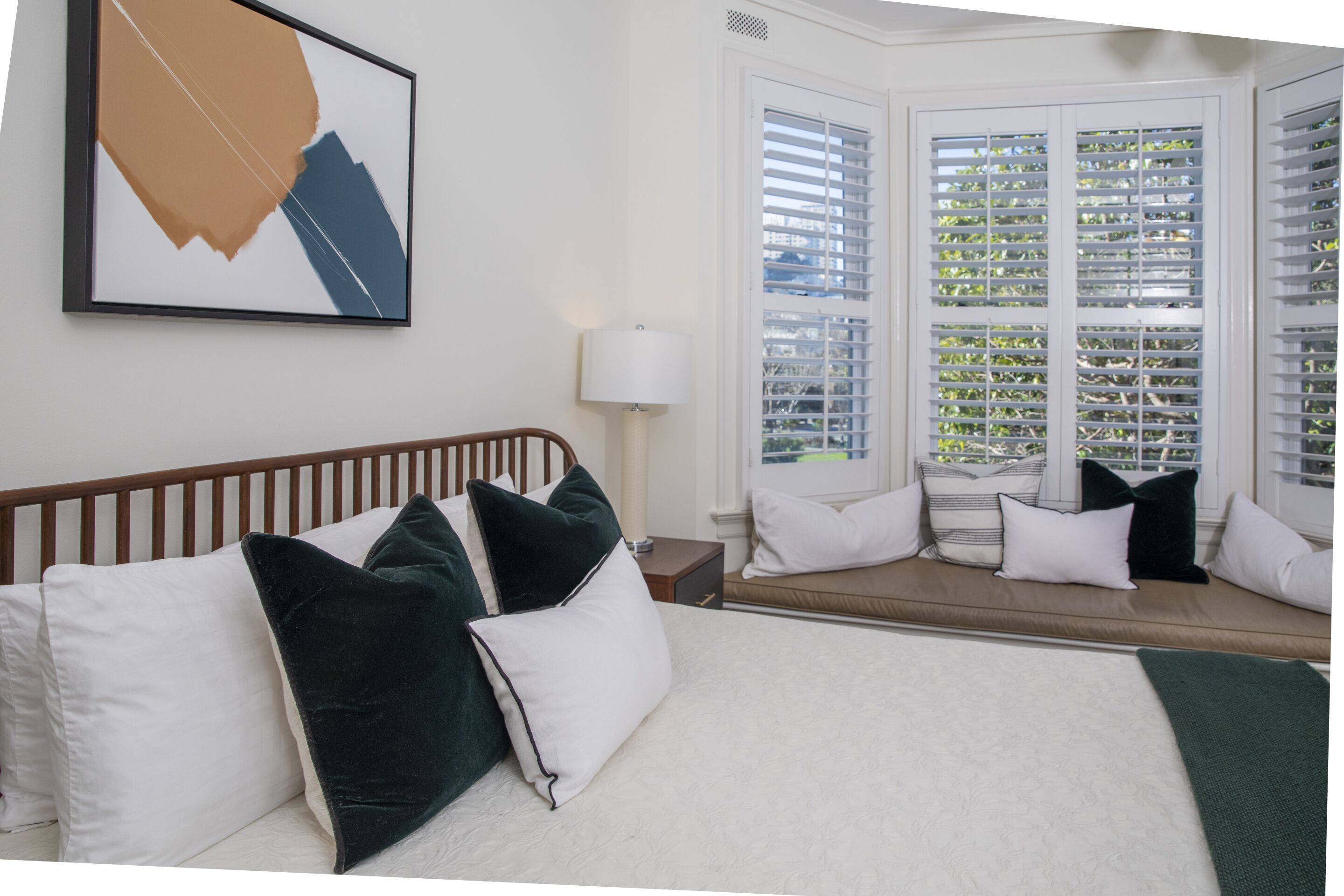 A hotel room with a bed, a window with blinds, a bedside table, and a wall-mounted artwork.