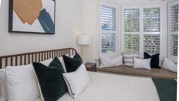 A hotel room with a bed, a window with blinds, a bedside table, and a wall-mounted artwork.