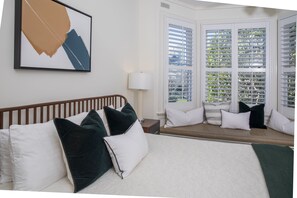 A hotel room with a bed, a window with blinds, a bedside table, and a wall-mounted artwork.