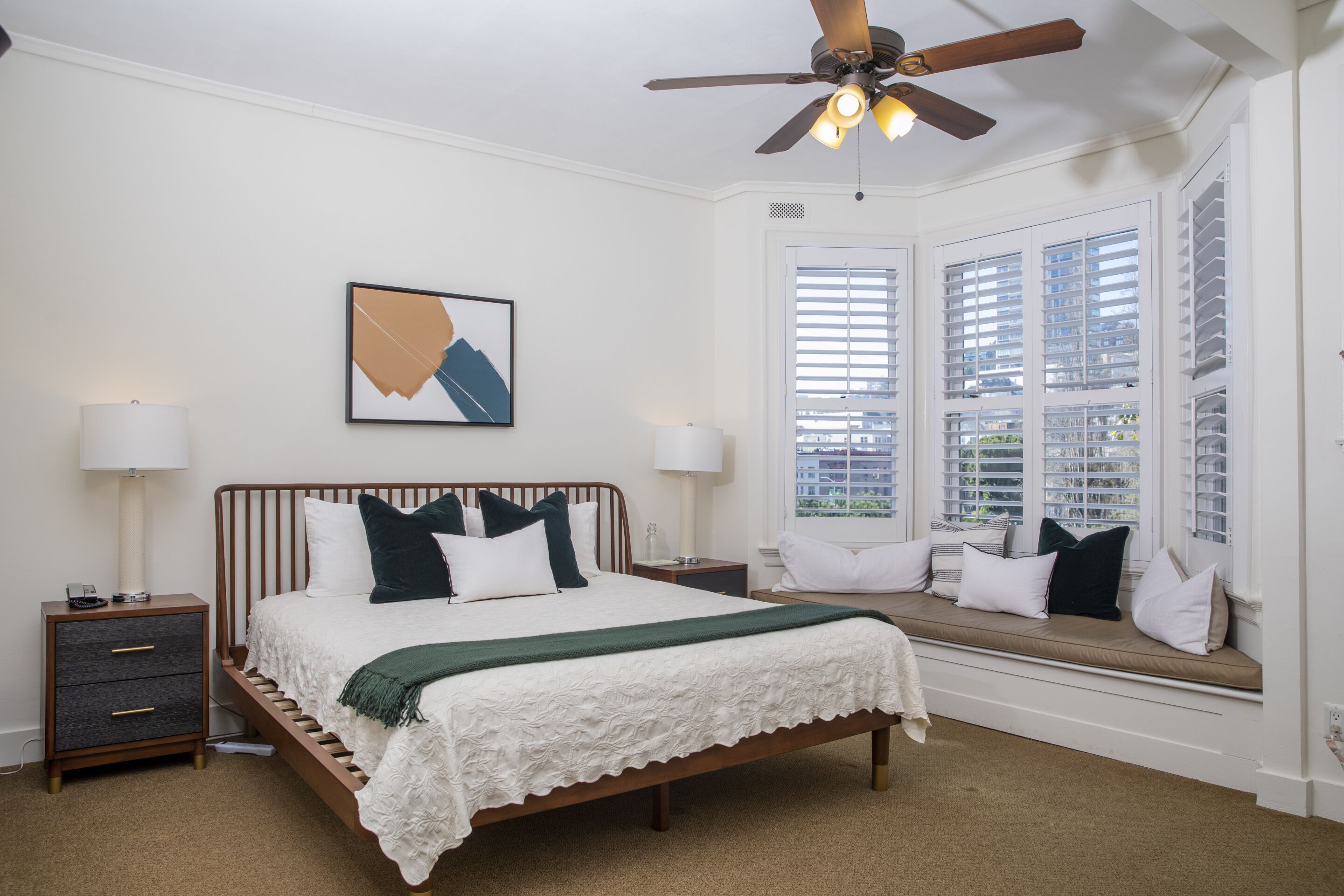A bedroom with a bed, bedside tables, a ceiling fan, a window with blinds, and a view of the outside.
