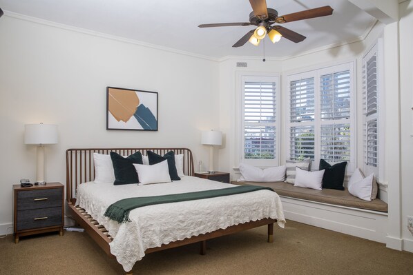 A bedroom with a bed, bedside tables, a ceiling fan, a window with blinds, and a view of the outside.