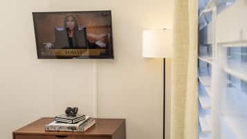 A television mounted on a wall displaying a news broadcast, a wooden nightstand with books and a decorative object, and a lamp beside the wall.