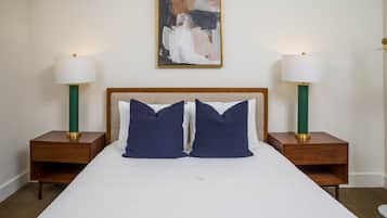 A neatly made bed with two pillows, flanked by two bedside tables with lamps, in a room with a plain wall and a single abstract painting.