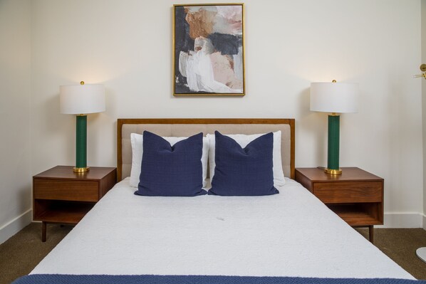A neatly made bed with two pillows, flanked by two bedside tables with lamps, in a room with a plain wall and a single abstract painting.