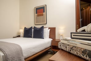 A neatly made bed with a white comforter and dark pillows, a wooden nightstand with a lamp, and a stack of books on the nightstand. - Standard Queen Room with Ensuite Bathroom (San Francisco)