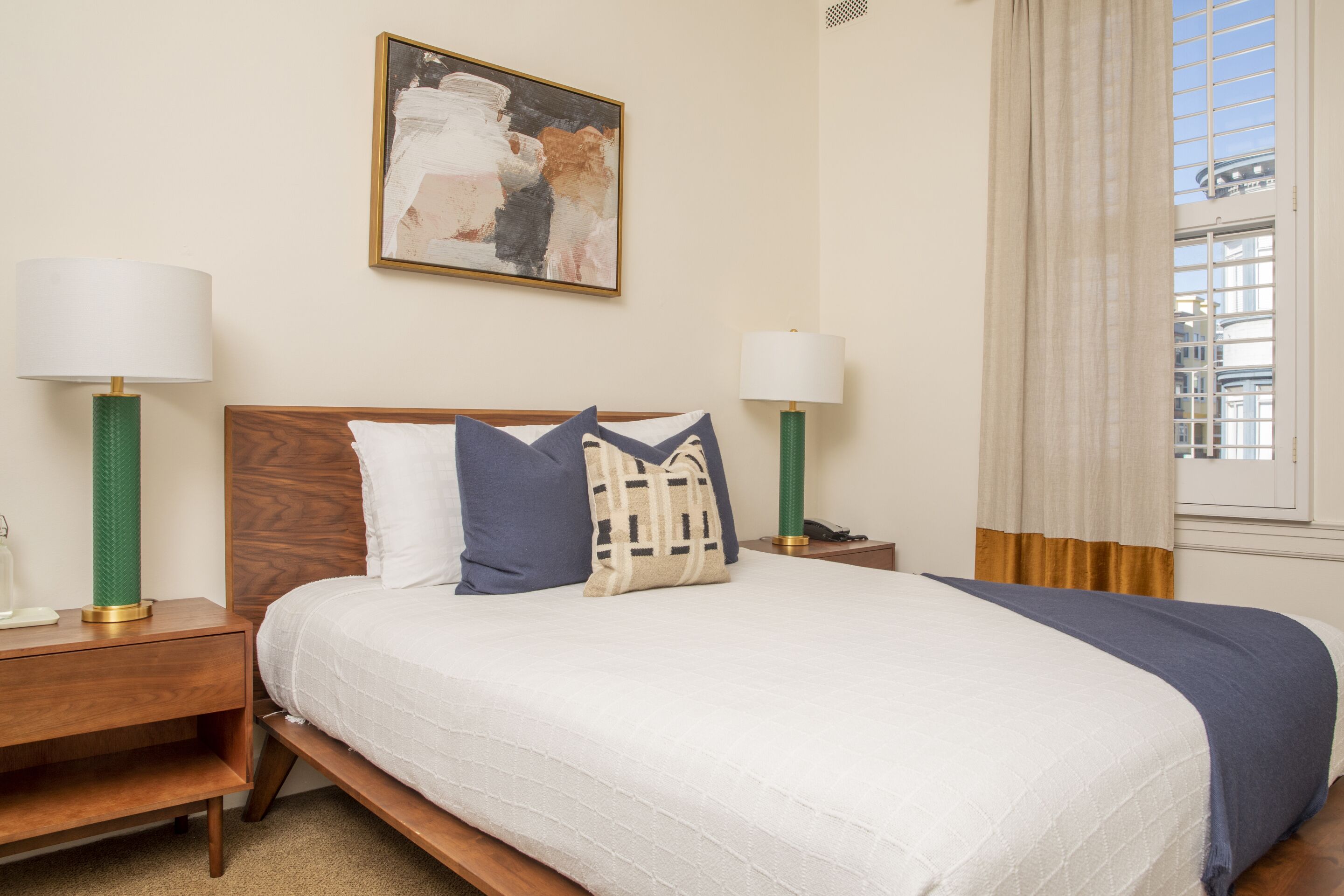 A neatly made bed with a white and blue bedspread, two bedside lamps, a wooden nightstand, and a painting on the wall.