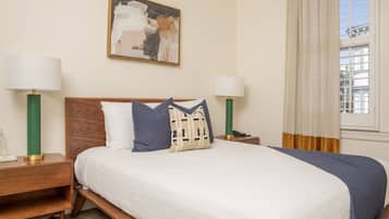 A neatly made bed with a white and blue bedspread, two bedside lamps, a wooden nightstand, and a painting on the wall.