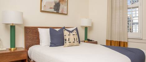 A neatly made bed with a white and blue bedspread, two bedside lamps, a wooden nightstand, and a painting on the wall.
