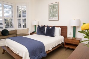 Un dormitorio ordenado con cama, mesitas de noche, un banco y una ventana con persianas.
