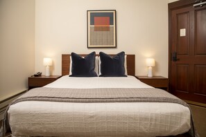Ein ordentlich bezogenes Bett mit einer gesteppten Decke, zwei Nachttischlampen, einem gerahmten Kunstwerk darüber und einer Holztür zur rechten Seite.