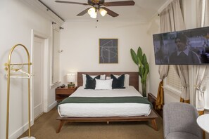 Une chambre d’hôtel comprenant un lit, une télévision, un ventilateur de plafond, une table de chevet, une lampe de chevet, une chaise et une plante.