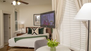 Een hotelkamer met een bed, een aan de muur bevestigde televisie, een nachtkastje met een lamp, een raam met zonwering, en een kleine ronde tafel met een bloevas.