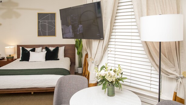 Een hotelkamer met een bed, een aan de muur bevestigde televisie, een ronde tafel met een bloevas en een lamp.