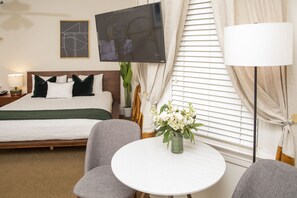 Une chambre d’hôtel avec un lit, un téléviseur fixé au mur, une table ronde ornée d’un vase de fleurs et une lampe.