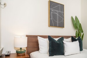 A neatly made bed with white and dark green pillows, a bedside lamp, a framed abstract artwork, and a potted plant.