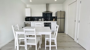 A compact kitchen with white cabinetry, a stainless steel range hood, a refrigerator, and a dining table with chairs. - Ohope Villas (Ohope)