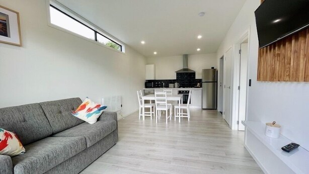 A modern living room with a grey sofa, a dining area with white chairs, a kitchen with a stove and refrigerator, and a TV mounted on the wall. - Ohope Villas (Ohope)