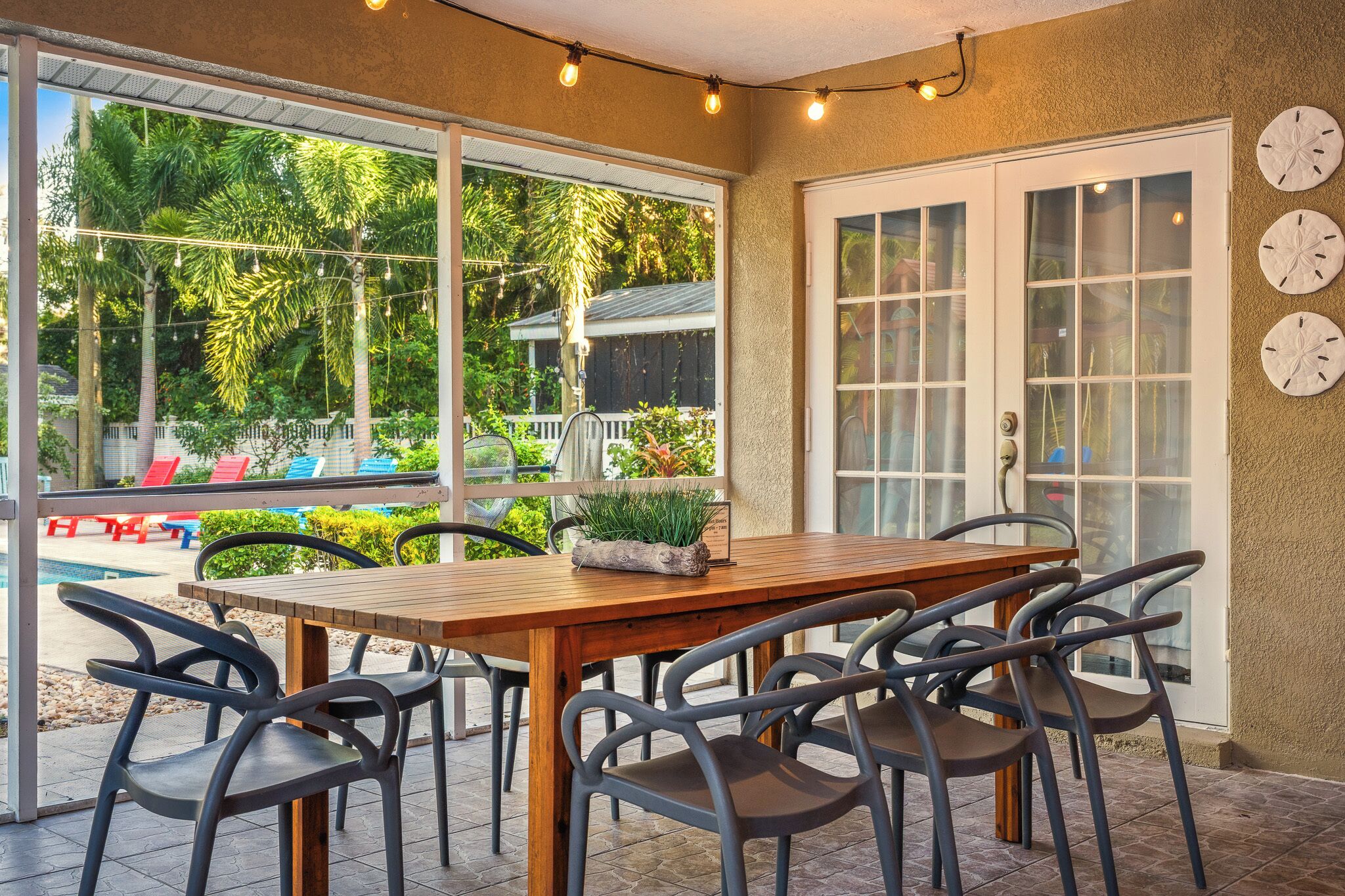 A patio area with a wooden table and chairs, large windows, and a view of a pool and greenery.