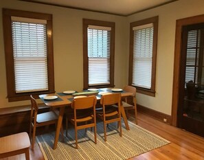 A dining room with a wooden table, four chairs, and three windows with blinds.