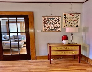 A room with a wooden floor, a sofa, a small wooden side table with a lamp, and two framed botanical prints on the wall.