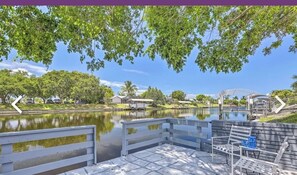 Um deck à beira do lago com um banco e uma mesa pequena, rodeado por árvores e um céu claro.