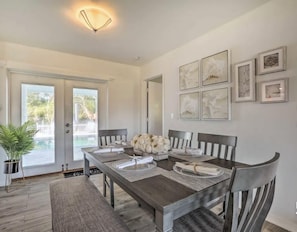 A dining area with a wooden table set for four, surrounded by chairs, with a potted plant to the left and framed artwork on the walls.