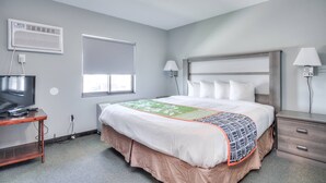 A hotel room with a bed, a TV on a stand, a window, and a bedside lamp. - Suite 218 - 1BR Apartment, King Bed, Kitchen, 2nd (Rapid City)