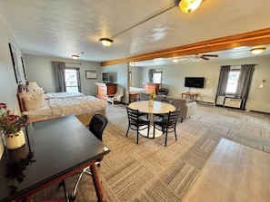 A hotel room with a bed, a desk, a dining table, a TV, and a ceiling fan.
