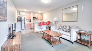 A modern living room with a sofa, coffee table, and red chairs. A kitchen with pink cabinets and a refrigerator is visible in the background.