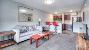 A living room with a sofa, coffee table, and red bar stools. A kitchen with cabinets and appliances is visible in the background.