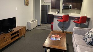 A living room with a TV, a coffee table with books, a sofa, and a kitchen area with a microwave and red bar stools. - 1BR with 2 beds and a kitchen on the main floor (Rapid City)