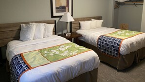 A hotel room with two beds, a nightstand with a lamp, a framed picture on the wall, and a shelf with folded clothes. - 1BR with 2 beds and a kitchen on the main floor (Rapid City)