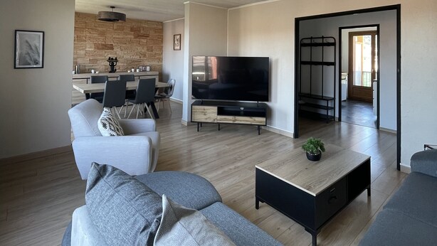 A modern living room with a sofa, coffee table, and TV. There is a dining area with chairs and a table in the background.
