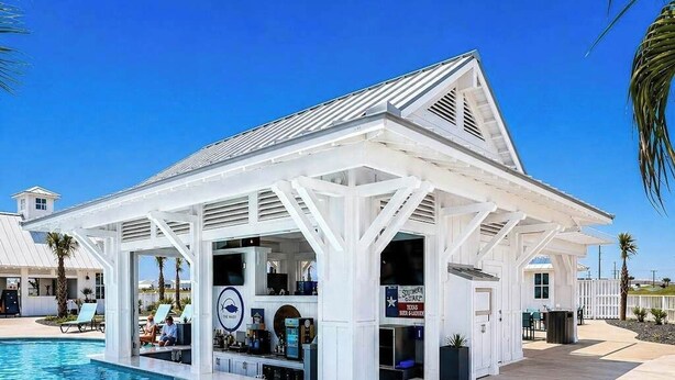 Un bar situé au bord de la piscine, avec une façade blanche, une piscine à l’eau d’un bleu limpide et un palmier en arrière-plan.