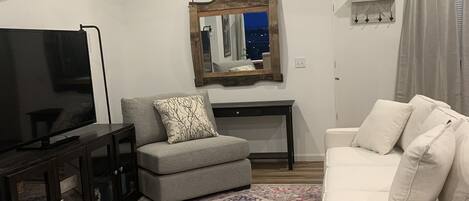 A living room with a gray sofa, a gray armchair, a television on a cabinet, a ceiling fan, a patterned rug, and a mirror on the wall.