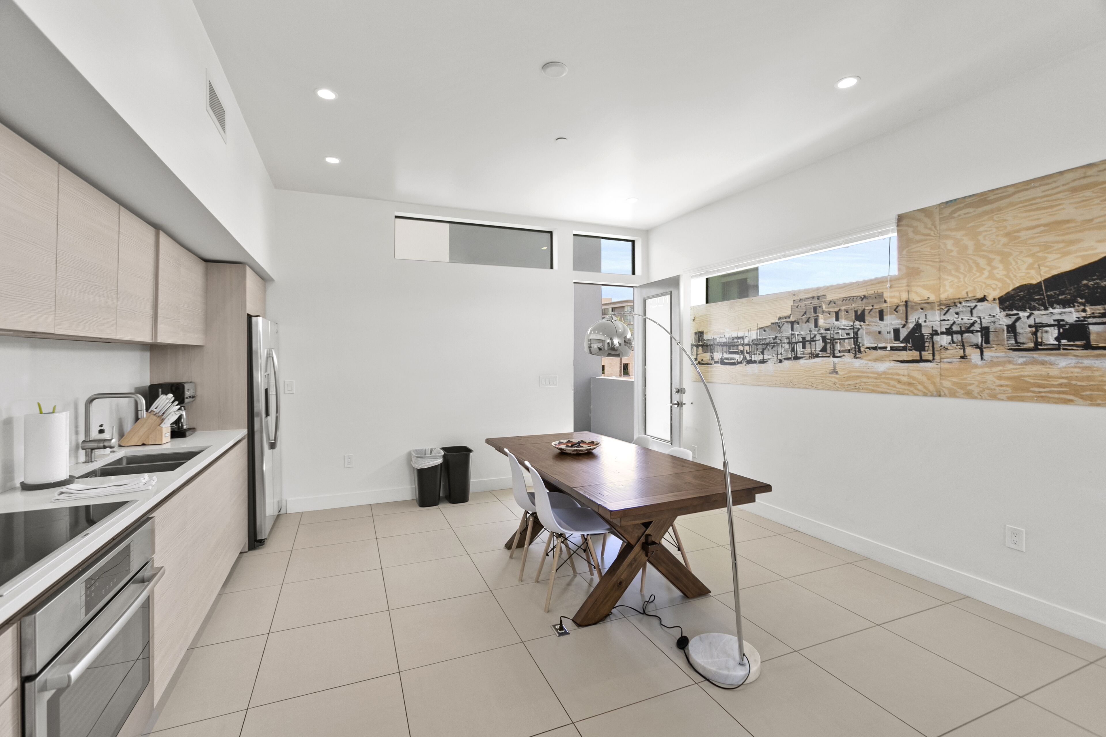 A modern kitchen with a wooden dining table, white cabinets, and a large window.
