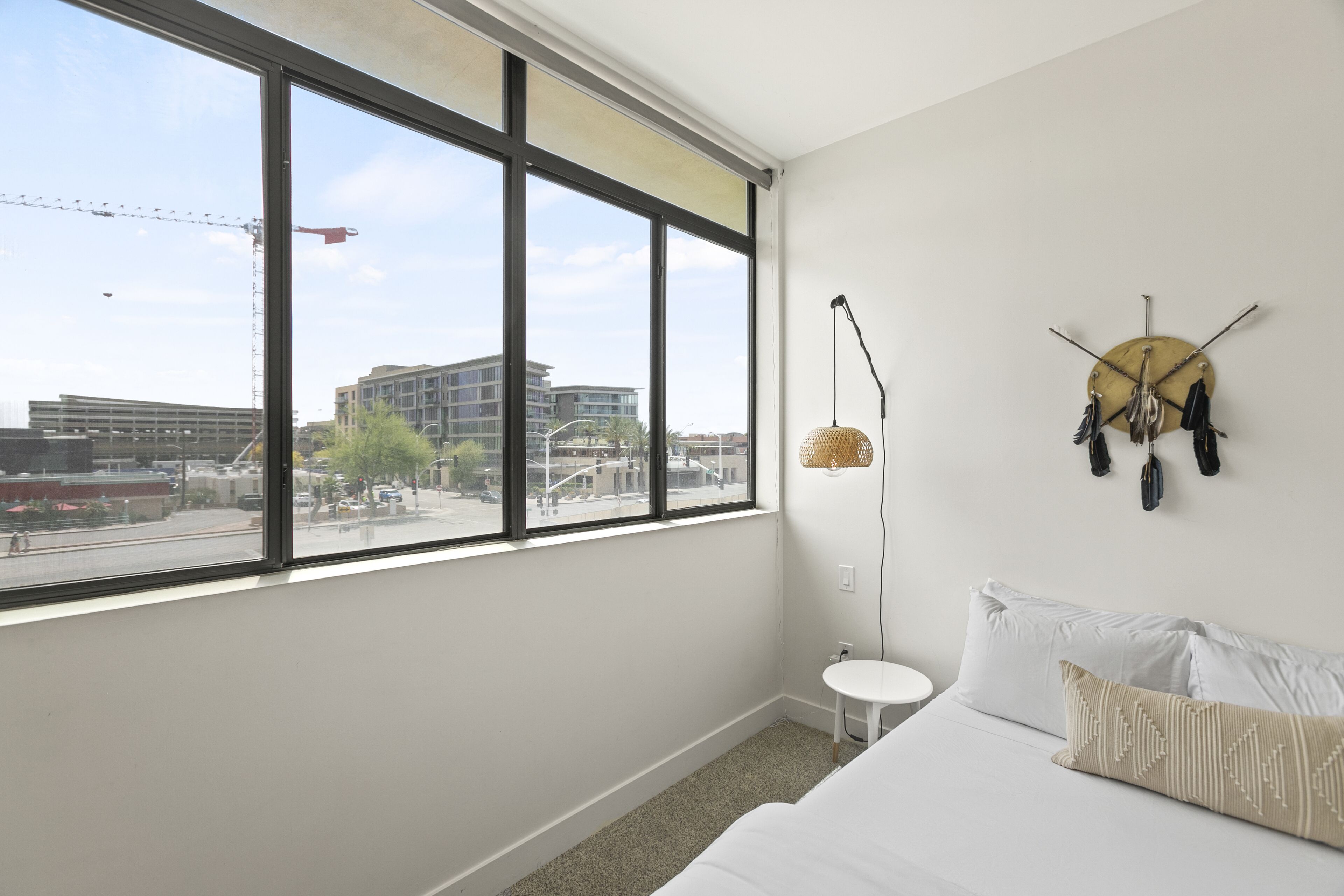 A modern hotel room with a large window, a bed with white linens, a small white side table, and a wall-mounted dreamcatcher.