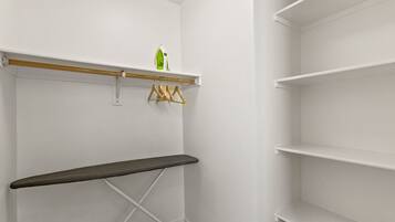 A white closet with a wooden bench, a hanging rack with clothes, and a shelf with a green bottle.