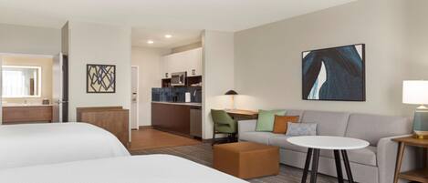 A hotel room with two beds, a sofa, a small table, a lamp, and a kitchen area in the background.