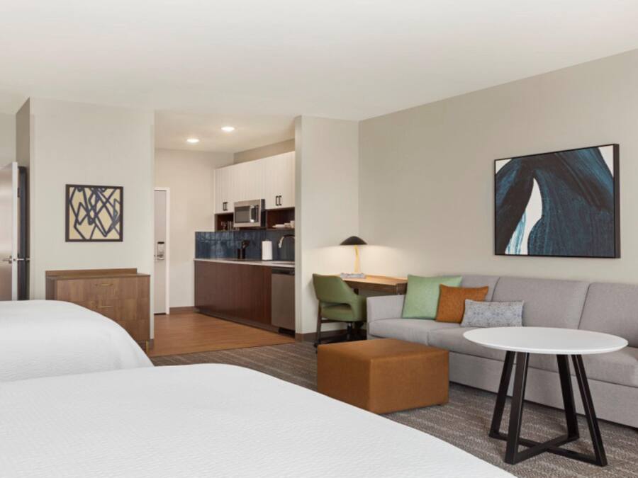 A hotel room with two beds, a sofa, a small table, a lamp, and a kitchen area in the background.