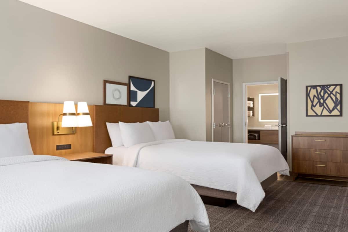 A hotel room with two beds, a nightstand with lamps, a door, a bathroom mirror, and a dresser.