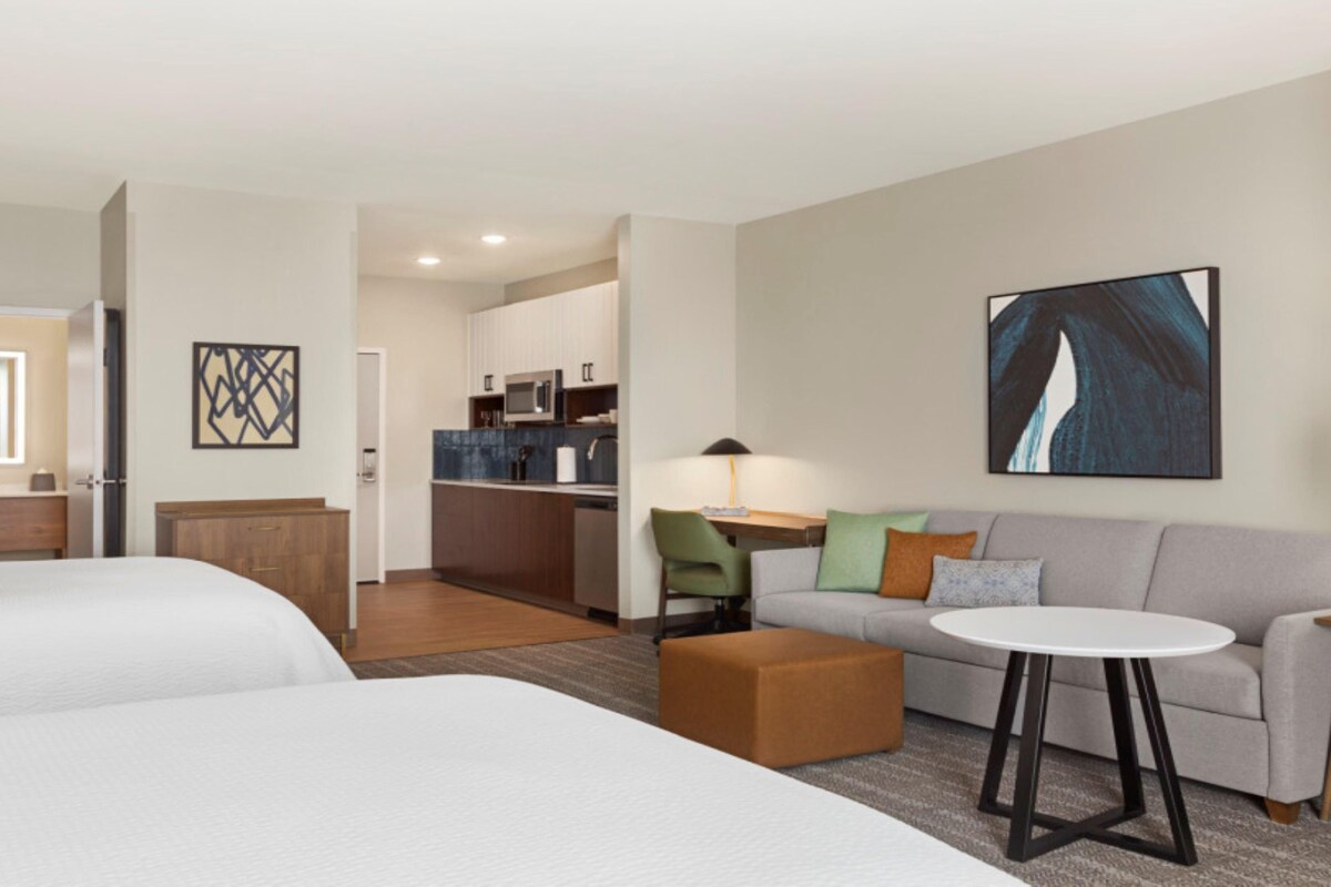 A hotel room with two beds, a sofa, a small table, a lamp, and a kitchen area in the background.