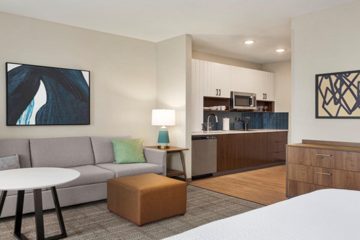 A modern hotel room with a grey sofa, a small round table, a brown ottoman, a bed, a kitchenette, and a large abstract painting on the wall.