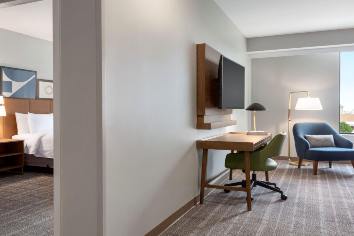 A hotel room with a bed, desk, chair, TV, and a view of the city.
