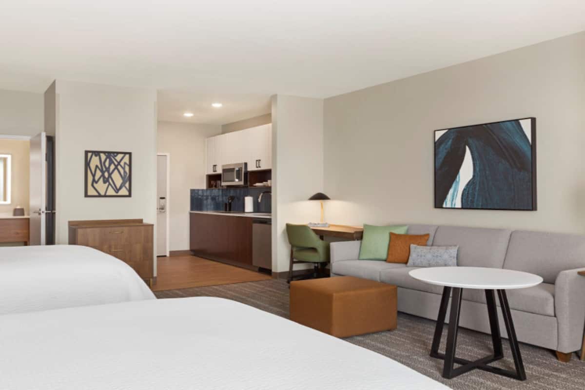 A hotel room with two beds, a sofa, a small table, a lamp, and a kitchen area in the background.